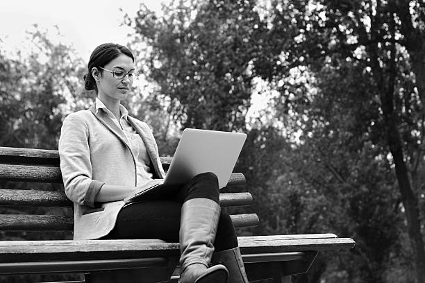 Young woman using laptop sitting on bench in park BnW