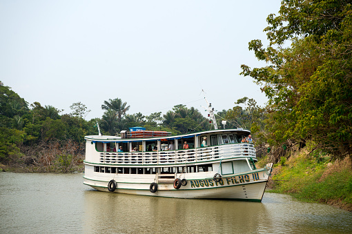 Santarem, Brazil - December 2, 2015: white cruise boat with tourists on deck floating on dirty green river water near trees on coast on natural background
