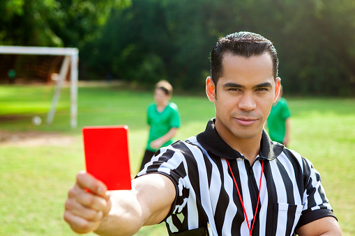 Serious mid adult Hispanic referee looks at the camera while holding out a red penalty card during a youth soccer game. He is wearing a referee uniform. Soccer players are in the background.