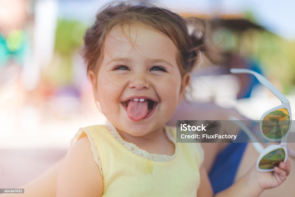 portrait of baby girl with sunglasses on a beach Baby girl posing on a beach with white sunglasses gold mirror. Enjoying sun and a beautiful day - wearing yellow shirt without sleeves . She is exults, bragging, mocking Baby - Human Age Stock Photo