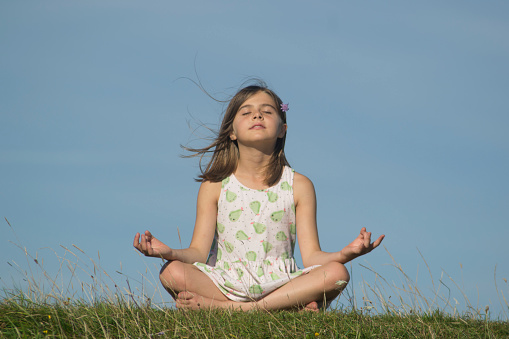 A young girl sits in the lotus position meditating in the long grass on a warm summers day. 