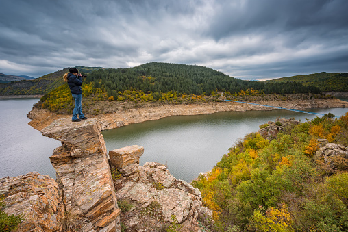 A woman on the top of a rock taking a picture of an autumn forest at Kardzhali dam, Bulgaria