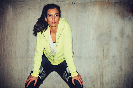 Portrait of an professional female runner against concrete wall, lighted with flash. Holding hands on knees.