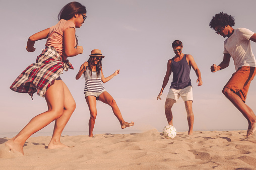 Group of cheerful young people playing with soccer ball on the beach with sea in the background