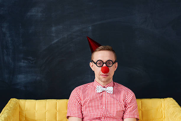 Boring young man wearing clowns nose, glasses and party hat sitting on sofa