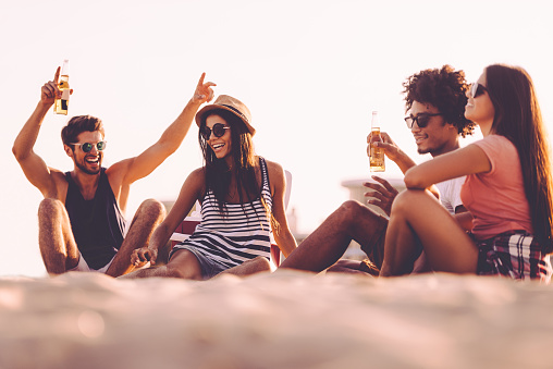 Cheerful young people spending nice time together while sitting on the beach and drinking beer