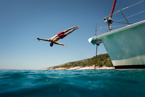 Muscular mid adult man diving into the sea water from deck of anchored yacht or sailboat. He is wearing red swimwear which makes gread contrast with surrounding colors. Photo taken from sea surface level.
