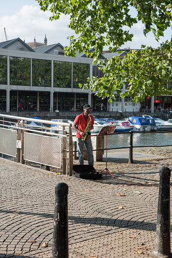 Bristol, UK - September 14, 2016: a senior black man playing saxophone on Bristol's quayside. He has set up his music stand under a tree by Pero's bridge - a bridge commemorating a famous freed black slave in Georgian Bristol - and the boats on Bristol's floating harbour can be seen in the background.