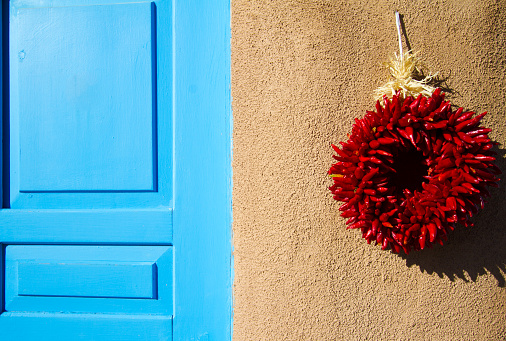 Santa Fe style: A red chili pepper ristra wreath hangs on an adobe wall next to a blue shutter/door. Ristras on doors are not just decorative in New Mexico, but are believed to ward off evil.