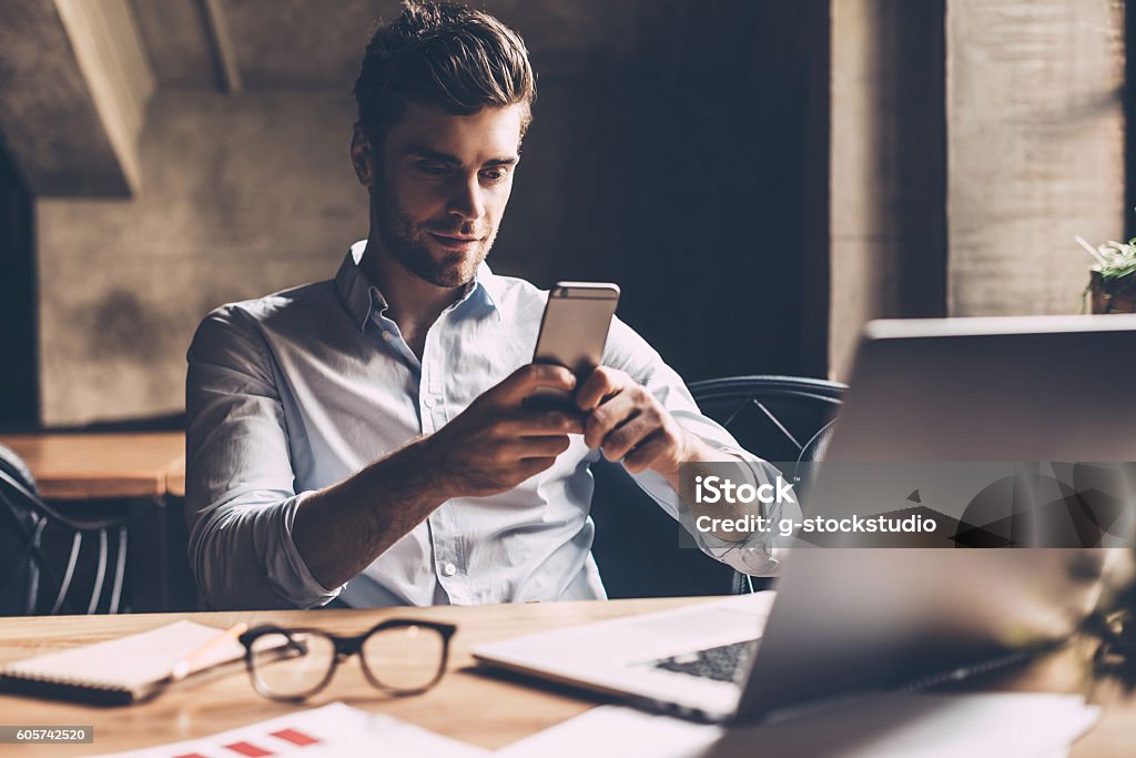 Texting to colleague. Confident young man in smart casual wear holding smart phone and looking at it while sitting at his working place in office Smart Phone Stock Photo