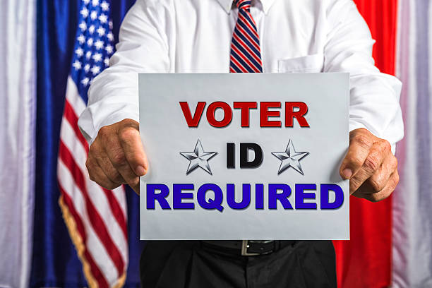 Businessman holding a sign that reads Voter ID Required A middle-aged Caucasian man wearing a white button down shirt, a red white and blue tie and black slacks, holding a sign that reads " Voter ID Required" while standing in a Polling station with red, white and blue drapes and an American flag in the background. voter id stock pictures, royalty-free photos & images