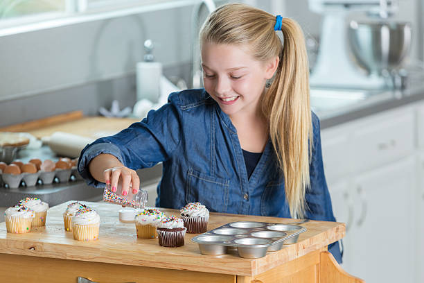 istock Young blonde girl decorates cupcake with sprinkles in kitchen 605745144