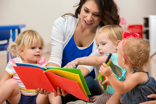 Hispanic young adult female teacher reading a colorful book to cute blonde toddler girls in her preschool classroom. They are sitting on the floor and interested in the book.