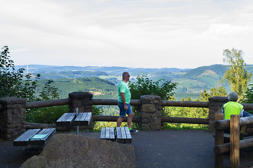 Lennestadt, Germany - September 11, 2016: Capture of some people at look out Hohe Bracht in Sauerland in summertime. Afternoon shot. A man is standing near wooden fence and railing. Bikers are sitting in right area.