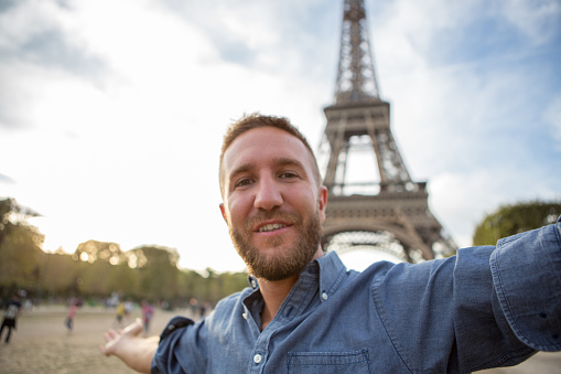 Young cheerful man near the Eiffel tower in Paris-France take a selfie portrait using a mobile phone. Beautiful summer day.