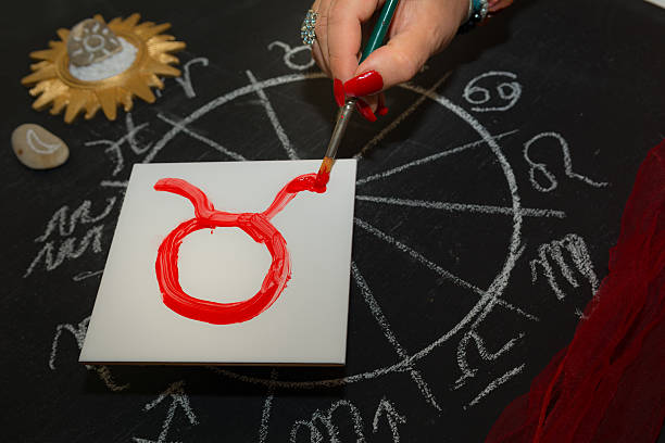 Female astrologer draws taurus zodiac sign on white tile Mature woman astrologer with red nails draws a zodiac sign on a white tile above the full zodiac drawn on a blackboard with white chalk in a circle. All drawings made by contributor. Western astrology is  based on the construction of a horoscope for an exact moment, such as a person's birth. It uses the tropical zodiac, which is aligned to the equinoctial points. taurus photos stock pictures, royalty-free photos & images