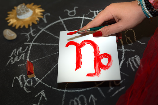 Mature woman astrologer with red nails draws a zodiac sign on a white tile above the full zodiac drawn on a blackboard with white chalk in a circle. All drawings made by contributor. Western astrology is  based on the construction of a horoscope for an exact moment, such as a person's birth. It uses the tropical zodiac, which is aligned to the equinoctial points.
