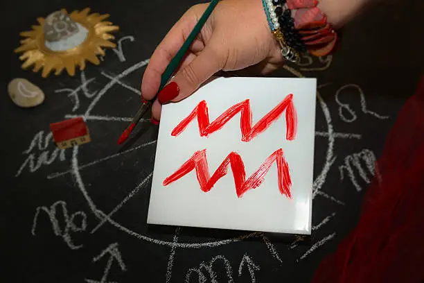 Mature woman astrologer with red nails draws a zodiac sign on a white tile above the full zodiac drawn on a blackboard with white chalk in a circle. All drawings made by contributor. Western astrology is  based on the construction of a horoscope for an exact moment, such as a person's birth. It uses the tropical zodiac, which is aligned to the equinoctial points.