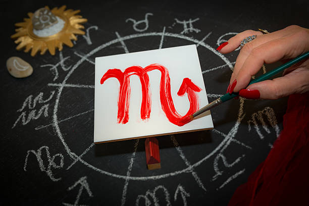 Mature woman astrologer with red nails draws a zodiac sign on a white tile above the full zodiac drawn on a blackboard with white chalk in a circle. All drawings made by contributor. Western astrology is  based on the construction of a horoscope for an exact moment, such as a person's birth. It uses the tropical zodiac, which is aligned to the equinoctial points.