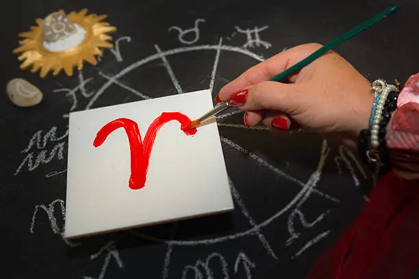 Mature woman astrologer with red nails draws a zodiac sign on a white tile above the full zodiac drawn on a blackboard with white chalk in a circle. All drawings made by contributor. Western astrology is  based on the construction of a horoscope for an exact moment, such as a person's birth. It uses the tropical zodiac, which is aligned to the equinoctial points.