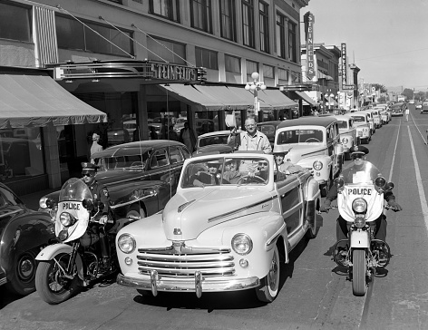 Tucson, Arizona, USA - November 4, 1947: 1947 Ford Super DeLuxe Sportsman “woodie” convertible leads a parade of new Ford cars in front of Steinfled’s department store along Stone Avenue in Tucson, Arizona. The event celebrates the opening of a new Monte Mansfield (in lead car) Ford showroom. Two motorcycle police on Harleys, escort the parade.