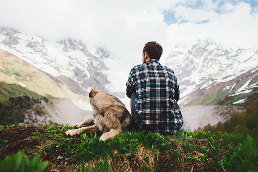 Young Caucasian man sitting on the grass with dog on the background of Caucasus mountains, Georgia 