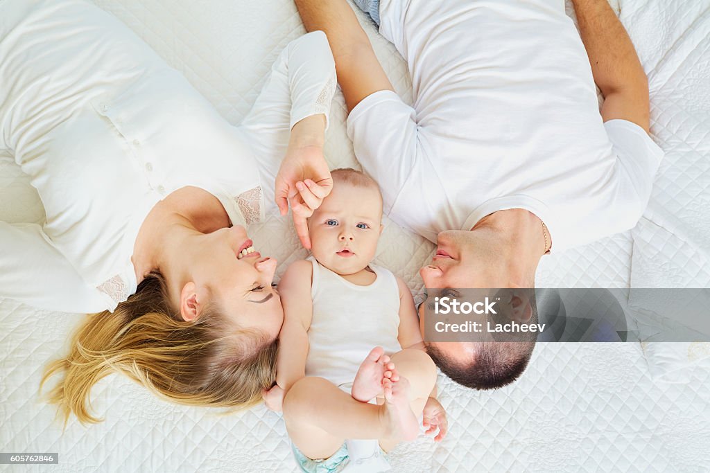 Concept of  happy family. Top view mom dad and baby The concept of a happy family. Top view of a mom, dad and baby boy lying on white bed in the room indors. Baby - Human Age Stock Photo