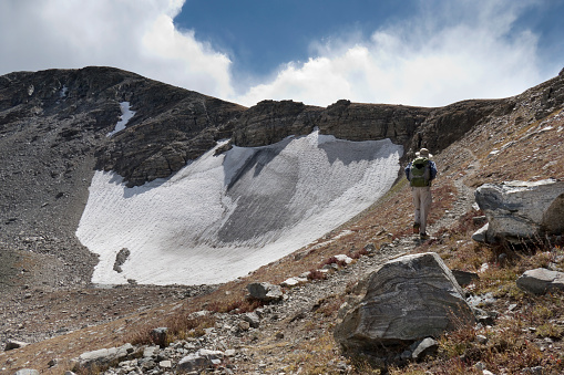 Looking towards the 12,304 foot high Sawtooth Mountain and the Rocky Mountain Continental Divide, a man hikes with a backpack past a snow field through the Indian Peaks Wilderness, Colorado.