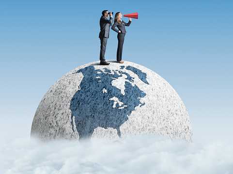 A businessman and a businesswoman stand on top of a globe that rests in the clouds.  He looks through a pair of binoculars as she shouts through a red megaphone as they try to communicate at a global level.