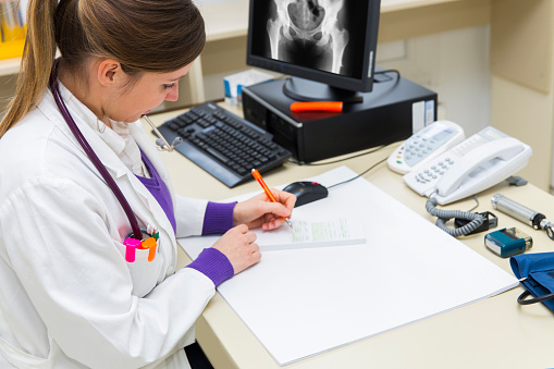 Pretty young woman doctor writing prescription...X-ray picture of human pelvis on monitor