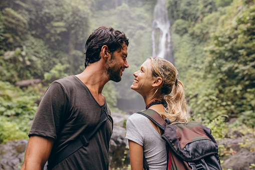 Affectionate young couple together on hike. Young man and woman with backpack looking at each other and smiling. Hiking in forest with waterfall in background.