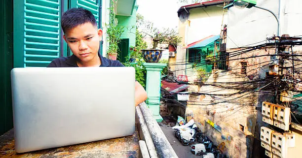 Panoramic view of a Young Vietnamese man working with laptop on a Hanoi balcony, with partial view of the street below.