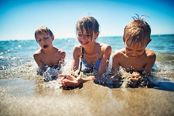 Kids having fun in sea lying on beach Little girl aged 10 with her brothers aged 7 are having fun lying on beach and being splashed by waves. Candid smile and laughter. Sunny summer day in Italy, Tuscany. girls laying on beach stock pictures, royalty-free photos & images