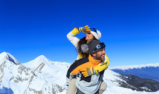 Cheerful young couple skiers having fun on the top of the snow mountain, in piggyback ride. Both wears warm clothes and gloves, with knit hats, ski gogglers. Snow mountain peak and clear sky on background.