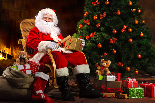 Santa Claus sitting in front of fireplace near Christmas tree with a bag full of presents and a wish list