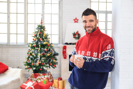 Smiling young man sitting at home and drinking coffee. Man with christmas pullover in red, blue and white leaning on brick column and holding a coffee cup. Looking at camera. On background decorated christmas tree and fireplace, presents on ground, a sofa and tall windows.