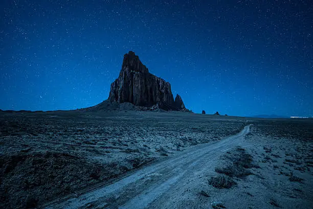 Night sky with many stars above Shiprock and a dirt road. Shiprock is a great volcanic rock mountain rising high above the high-desert plain of the Navajo Nation in New Mexico, USA