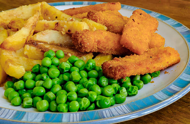 Small kid's meal - fish, chips, carrot and green peas. Colorful dinner on white wooden table with children's hands around. Plate captured from above (top view, flat lay).