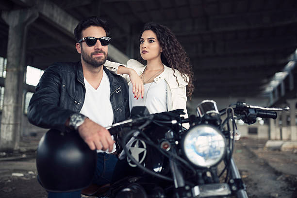 Bearded brutal man in sunglasses and leather jacket sitting on a motorcycle on the road in the forest with blured colorful background.