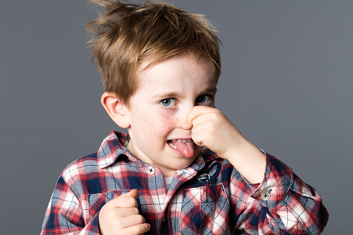 unhappy young boy pinching his nose for sign of bad odor, sticking out his tongue for humor and mischievous childhood, grey background