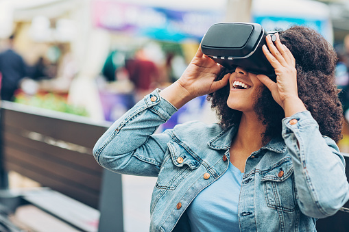 Young woman looking through a virtual reality headset and smiling outdoors on a bench, with copy space.