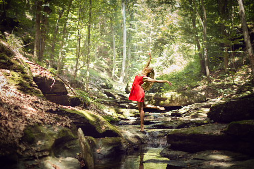 Beautiful ballerina, dancing ballet in green forest in the middle of the creek. She is wearing a short red dress and with bare feet standing one the rock in the stream and she spread hers arms as flying. She is happy and she is smiling.