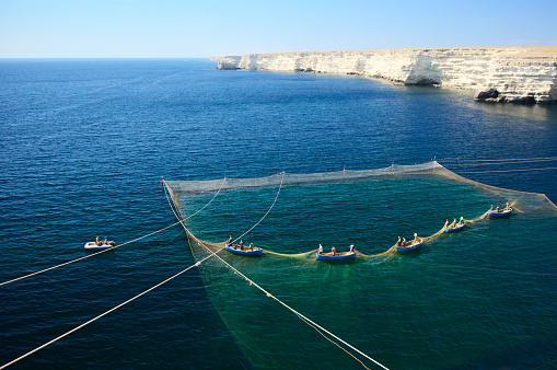 Old small fishing boats with fishermen drawing out nets in dark blue sea.