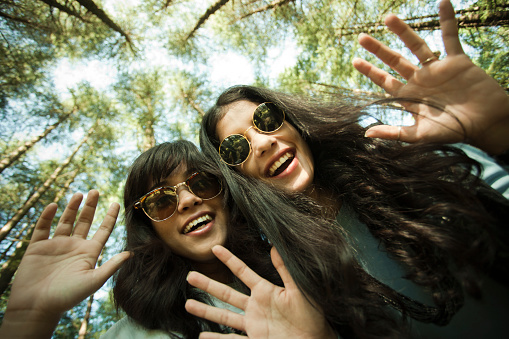 Outdoor worm eye view shot through wide angle lens showing tree top and sky in the background of two happy girls looking at camera with hand gesture and toothy smile, they are wearing sunglasses. Two people, low angle, waist up, horizontal composition with copy space.
