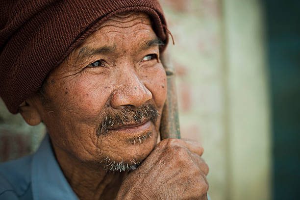 Outdoor image at day time of Asian serene senior peasant man sitting and thinking while looking away upward at sky by holding bamboo stick and resting his chin on hand. One person, head and shoulders, horizontal composition with selective focus and copy space.