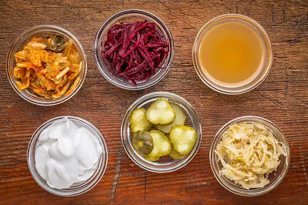 a set of fermented food great for gut health - top view of glass bowls against wood:  kimchi, red beets, apple cider vinegar, coconut milk yogurt, cucumber pickles, sauerkraut