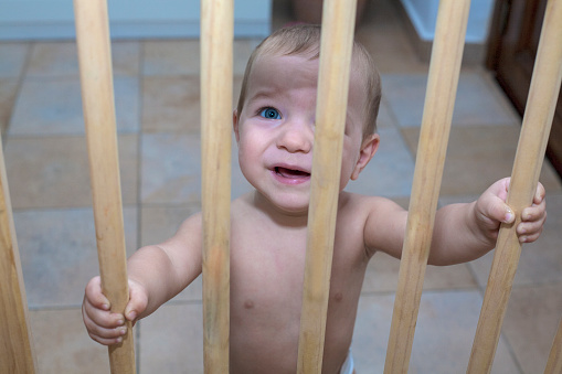 One year old baby boy behind the wooden safety gate of stairs. He is trying to go upstairs