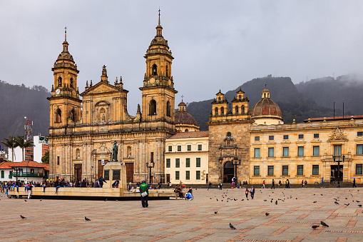 Bogota, Colombia - July 01, 2016: Looking from Plaza Bolivar, in the Andean Capital city of Bogota, in Colombia, South America, to the Eastern side of the Square. In full view is the Catedral Primada, the seat of the Roman Catholic Archbishop of Colombia. To the left of the image is the Casa del Florero and to the right, the Archbishop's chapel and a section of his palace. In the middle of the Square stand a statue of Simon Bolivar. The style of architecture is classical Spanish colonial. In the far background in the haze of the overcast, cloudy sky, the Andes Mountains can be seen.  People walk across the Square as they go about their daily work or explore the area. There are pigeons all over the square, and vendors sell grains to feed the pigeons. Photo shot in the afternoon sunlight, on a cloudy overcast day which works well to minimise harsh shadows; horizontal format. Copy space.