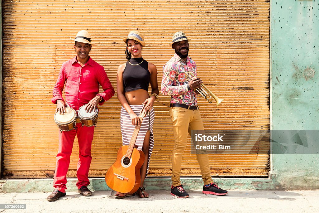Cuban Musical Trio Cuban musical band, the trio consisting of a well known musicians standing against a yellow closed shop shutter. Beautiful young woman standing in the middle, holding a guitar. The man on the left holding the small drums bongos, and a musician on the left holding a trumpet. Havana, Cuba Musician Stock Photo