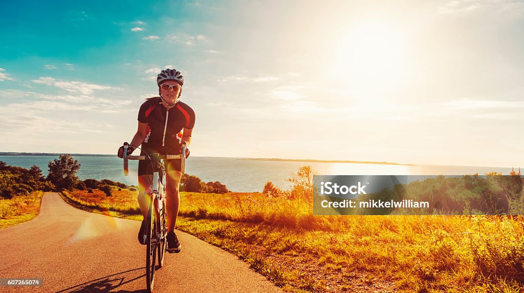 Cyclist rides her bike up a steep hill at sunset Close up of a female athlete who rides her bike up a steep hill. The sun shines as her feet hits the pedals and she drives the bicycle forward. In the background is the sun setting over the sea.  Cycling Stock Photo